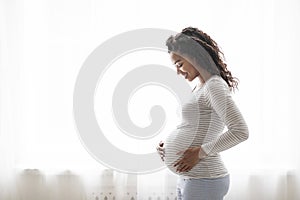 Beautiful pregnant black woman hugging tummy while standing near window at home