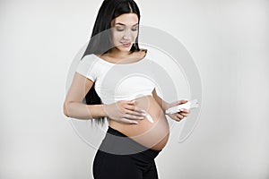 Beautiful pregnant brunette woman applying body lotion against strech marks on isolated white background