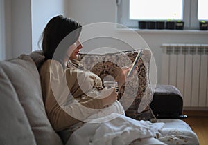 Beautiful pregnant Caucasian woman drinking tea while using a phone and sitting on a couch