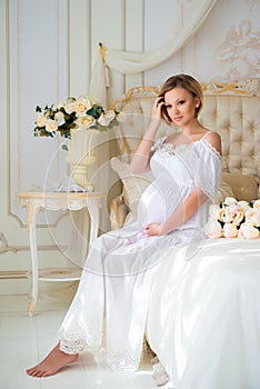 Beautiful pregnant girl in a home clothes sitting in the interior on a bed of roses