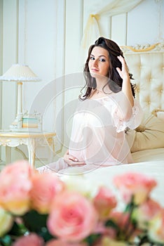 Beautiful pregnant girl in a lace negligee sitting on a bed of roses and touching hair.