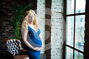 A beautiful pregnant girl stands near a window and looks at her tummy.