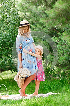 Beautiful pregnant girl in a straw hat is standing with a bouquet of summer flowers next to her little daughter and hugging in the