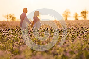 Beautiful pregnant mother and her husband walk together hand in hand outdoor, in the middle of a flowery field at sunset time. photo