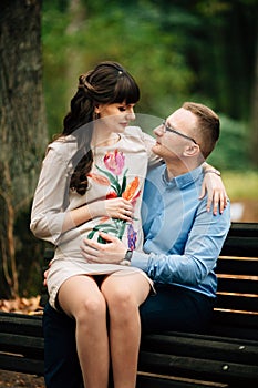 Beautiful pregnant stylish couple relaxing outside in the autumn park sitting on bench.