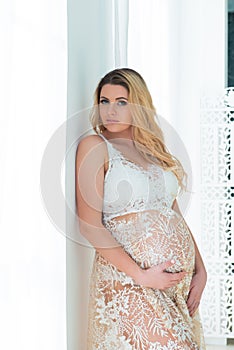 A beautiful pregnant woman in beautiful lace dress stands near a bright window. Feminine sexuality, femininity and beautiful
