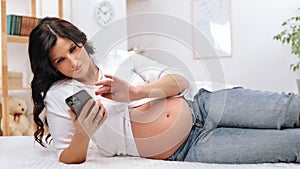 Beautiful pregnant woman with big tummy lying on bed and using smartphone, surfing internet, relax