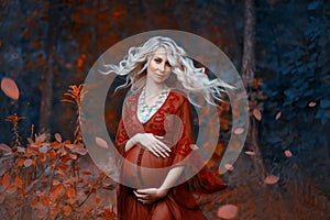 A beautiful pregnant woman with blond hair in long red dress and shiny necklace is standing in the forest, gently