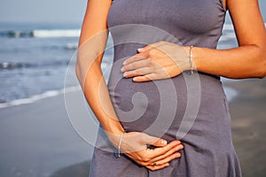 Beautiful pregnant woman in a blue grey tight dress is covered with hat on the beach touching her belly with love and