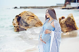 Beautiful pregnant woman in a blue tight dress is covered with a white striped plaid on the beach touching her belly