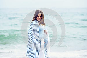 Beautiful pregnant woman in a blue tight dress is covered with a white striped plaid on the beach touching her belly