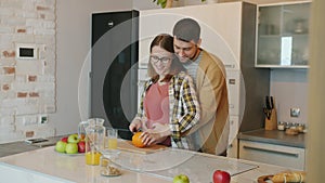 Beautiful pregnant woman cutting fruit when husband hugging her belly smiling in kitchen