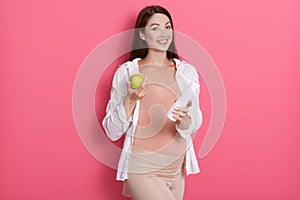 Beautiful pregnant woman with green apple and bottle of milk in hands, looking directly at camera while posing against pink wall,