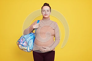 Beautiful pregnant woman with hair bun holds bag with baby`s things, prepares different items for newborn child going in maternit photo