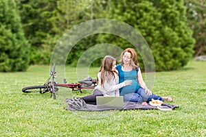 Beautiful pregnant woman and her daughter in a summer green park with laptop and bicycle