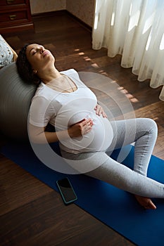 Beautiful pregnant woman with her eyes closed relaxing leaning on fitball, holding her belly, sitting on an exercise mat