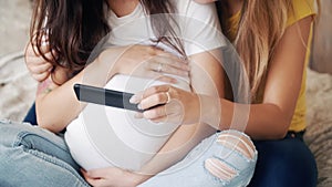 Beautiful pregnant woman holding and looking ultrasound photo newborn baby together with girlfriend on bed in bedroom