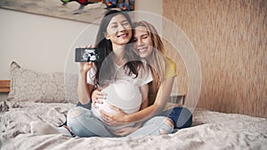 Beautiful pregnant woman holding and looking ultrasound photo newborn baby together with girlfriend on bed in bedroom