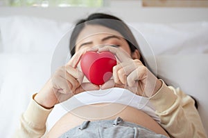 Beautiful pregnant woman lie down on bed and making hand heart on red heart ball at home. Pregnancy, motherhood, love and