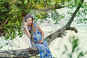 Beautiful pregnant woman mother to be in wreath and blue dress sitting on branch of tree near scenic river in sunset time in