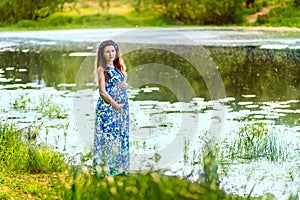 Beautiful pregnant woman mother to be in wreath and blue dress standing on bank of scenic river in sunset time in summer.