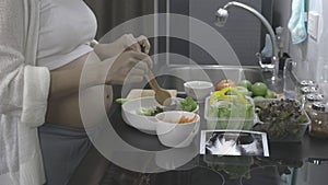 Beautiful pregnant woman preparing fresh vegetable salad in home kitchen.