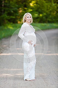 Beautiful pregnant woman in sheer long lacy white maternity dress looking dreamy on lonely road in forest