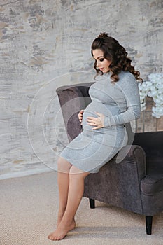 Beautiful pregnant woman sit on the armchair and put her hands to her belly. Woman dressed in elegant dress. Beautiful