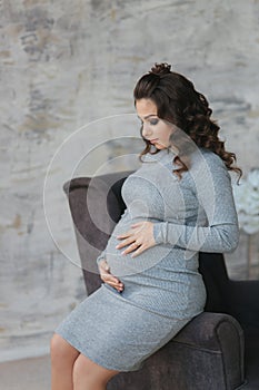 Beautiful pregnant woman sit on the armchair and put her hands to her belly. Woman dressed in elegant dress. Beautiful