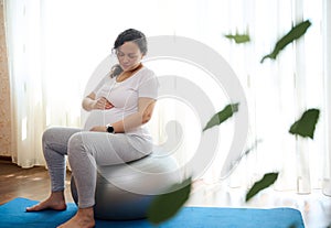 Beautiful pregnant woman sitting barefoot on fitball, holding hands on her belly, exercising in late pregnancy at home.