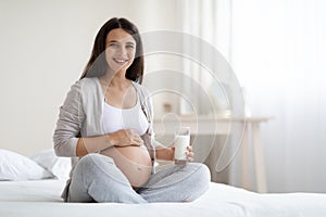 Beautiful pregnant woman sitting on bed, holding glass of milk