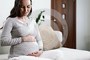 Beautiful pregnant woman sitting on couch with hands on pregnant belly, happy pregnancy