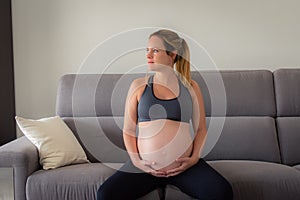 Beautiful pregnant woman sitting at sofa and keeping both hand on belly
