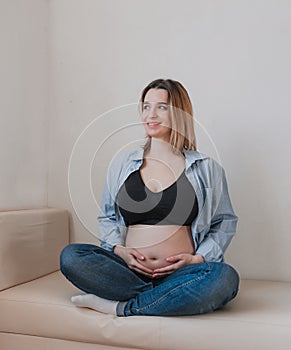 A beautiful pregnant woman is sitting on the sofa smiling and looking out the window.