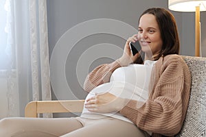 Beautiful pregnant woman sitting on sofa and using phone while calling to friend from home expressing positive emotions speaking