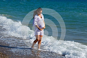 Beautiful pregnant woman standing on beach in light dress in the wind. Pleasure