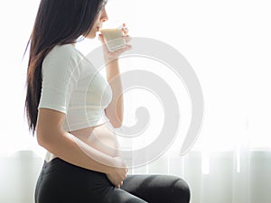 A beautiful pregnant woman standing, holding her stomach and drinking soy milk