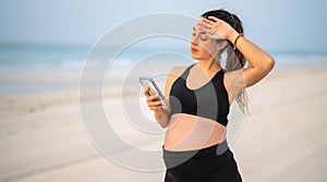 Beautiful pregnant woman standing over sandy beach seacoast looking at smart phone feeling tired holding hand on face.