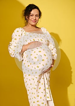 Beautiful pregnant woman in sundress, gently caresses her belly, smiles looking at camera, isolated on yellow background