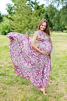 Beautiful pregnant woman walking barefoot in the park in long colorful dress with flying fabric. Happy maternity and fashion