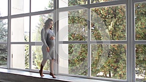 Beautiful pregnant woman walks in the house near a large window. She is dressed in a dress for pregnant women. Beautiful