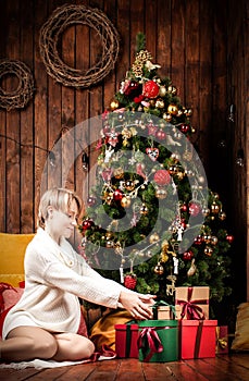 Beautiful pregnant woman wearing a warm sweater and socks, sitting near tha Christmas tree holding gift boxes
