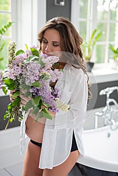 Beautiful pregnant woman in a white shirt in the bathroom near the window with a bouquet of lilac flowers. Soft selective focus