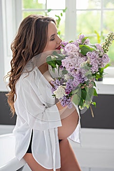Beautiful pregnant woman in a white shirt in the bathroom near the window with a bouquet of lilac flowers. Soft selective focus