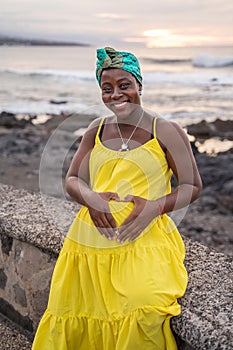 Beautiful pregnant woman in yellow fashionable dress posing on the beach, smiling and looking at the camera. Future motherhood