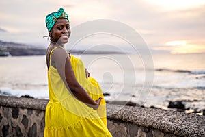 Beautiful pregnant woman in yellow fashionable dress posing on the beach, smiling and looking at the camera. Future motherhood