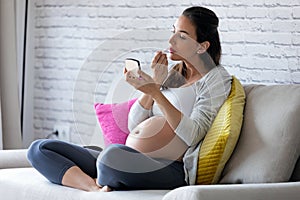 Beautiful pregnant young woman doing her makeup while sitting on sofa at home