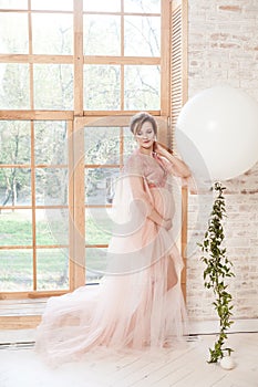 Beautiful pregnant young woman in pink dress standing near big natural window and touching her neck with hand