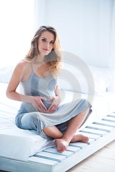 Beautiful pregnant young woman sits on a bed in a room and makes a heart on her tummy. Bright daylight sunlight from the windows