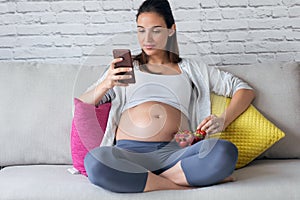 Beautiful pregnant young woman using her mobile phone while eating strawberries on sofa at home